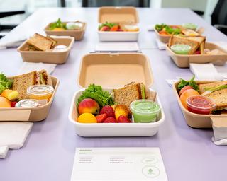 Individual packed lunch boxes containing sandwiches, fruit, snacks and drinks on an office table